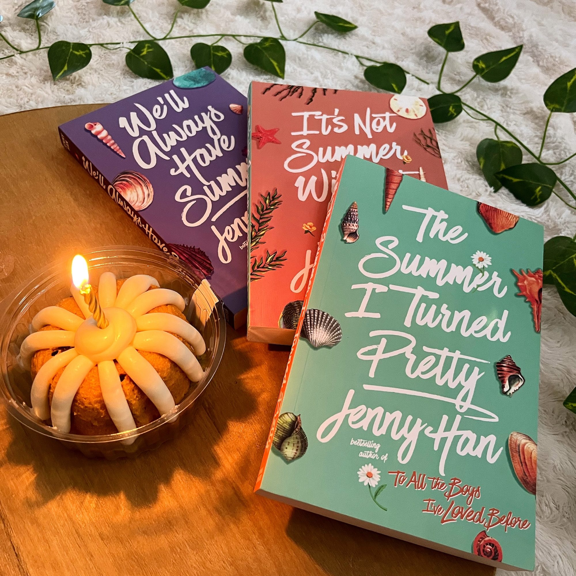 The The Summer I Turned Pretty trilogy by Jenny Han and a small bundt cake with a lit candle in its center sitting on a circular wooden tray and surrounded by a vine of green leaves.