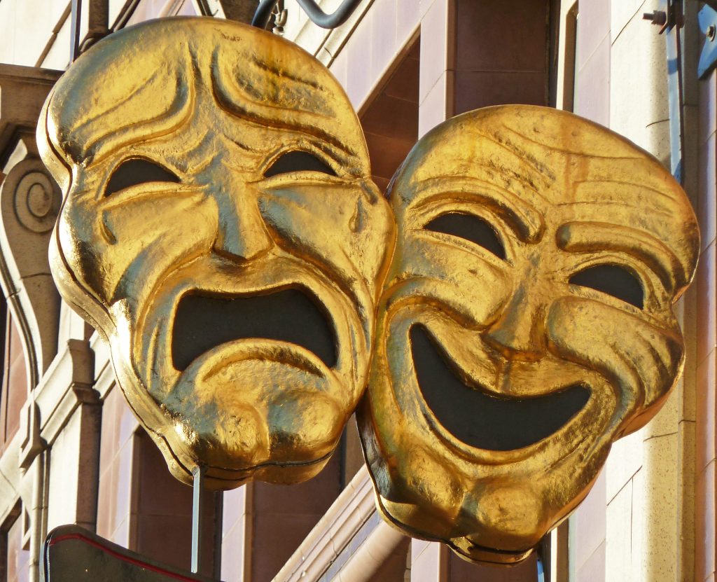 Two golden masks depicting tragedy and comedy on a hanging sign with a bulding in the bakground.