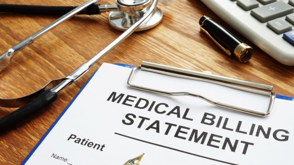 A brown table featuring a calculator, a stethoscope, and a medical billing statement attached to a blue clipboard.