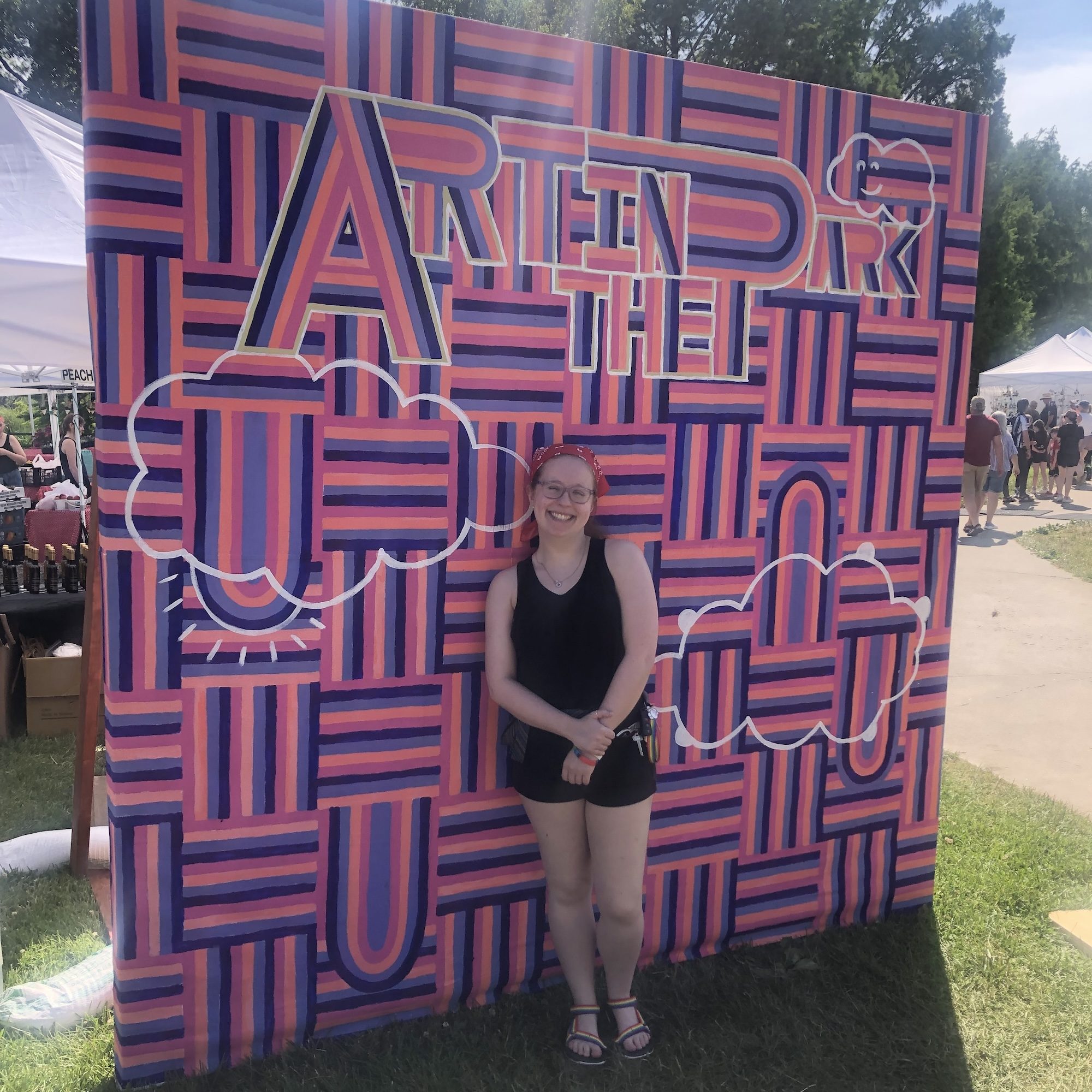 Haley standing in front of a pink and purple "Art in the Park" sign with white clouds on it.