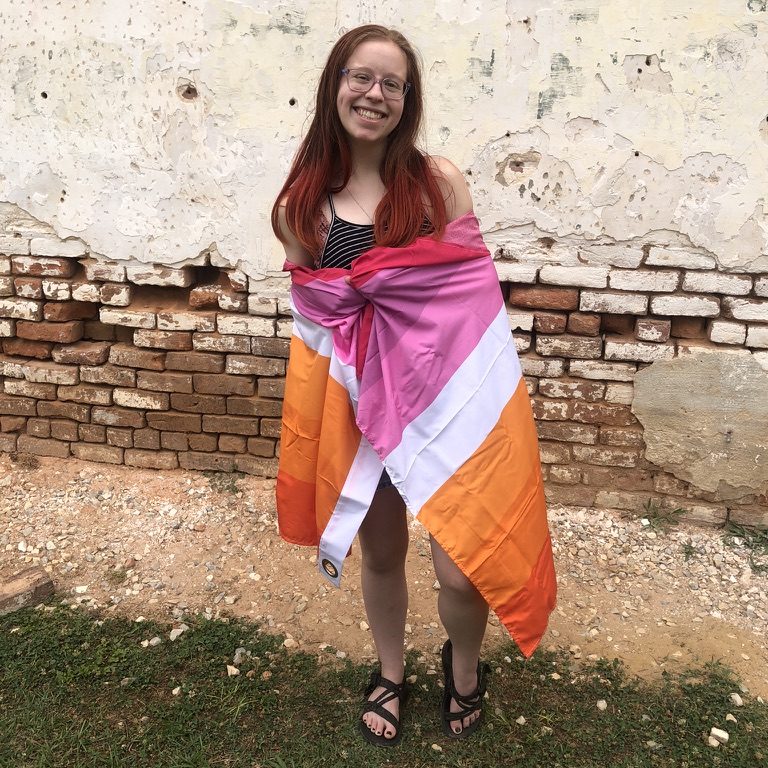 Haley standing in front of a brick wall with a lesbian flag around their shoulders.