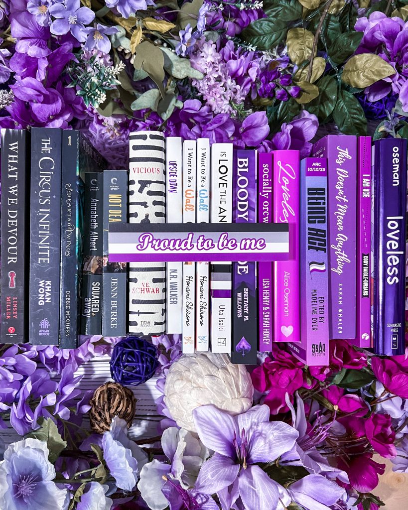 A variety of book spines arranged in a gradient from black to white to purple on a bed of purple flowers. There is an asexual pride flag bookmark featuring the phrase "proud to be me" sitting on top of the books.