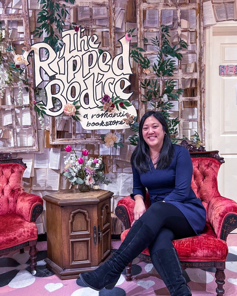 Rachel sitting in a red velvet chair in front of a wall of torn out book pages and a handmade sign that reads "The Ripped Bodice: A Romantic Bookstore"
