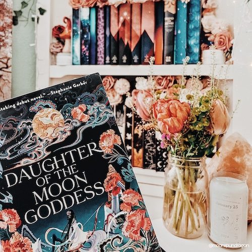 Book cover of "Daughter of the Moon Goddess" by Sue Lynn Tan, in front of a colorful bookshelf.