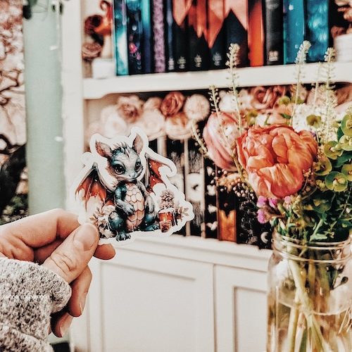 Dragon sticker held next to a flower vase, with a colorful bookshelf in the background.