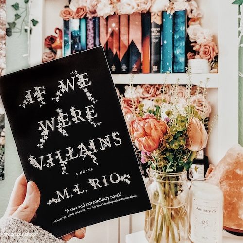 Book cover of "If We Were Liars" by M. L. Rio, held in front of a flower vase and colorful bookshelf.
