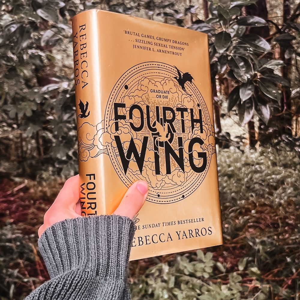 Hand holding Fourth Wing by Rebecca Yarros in front of a forest.