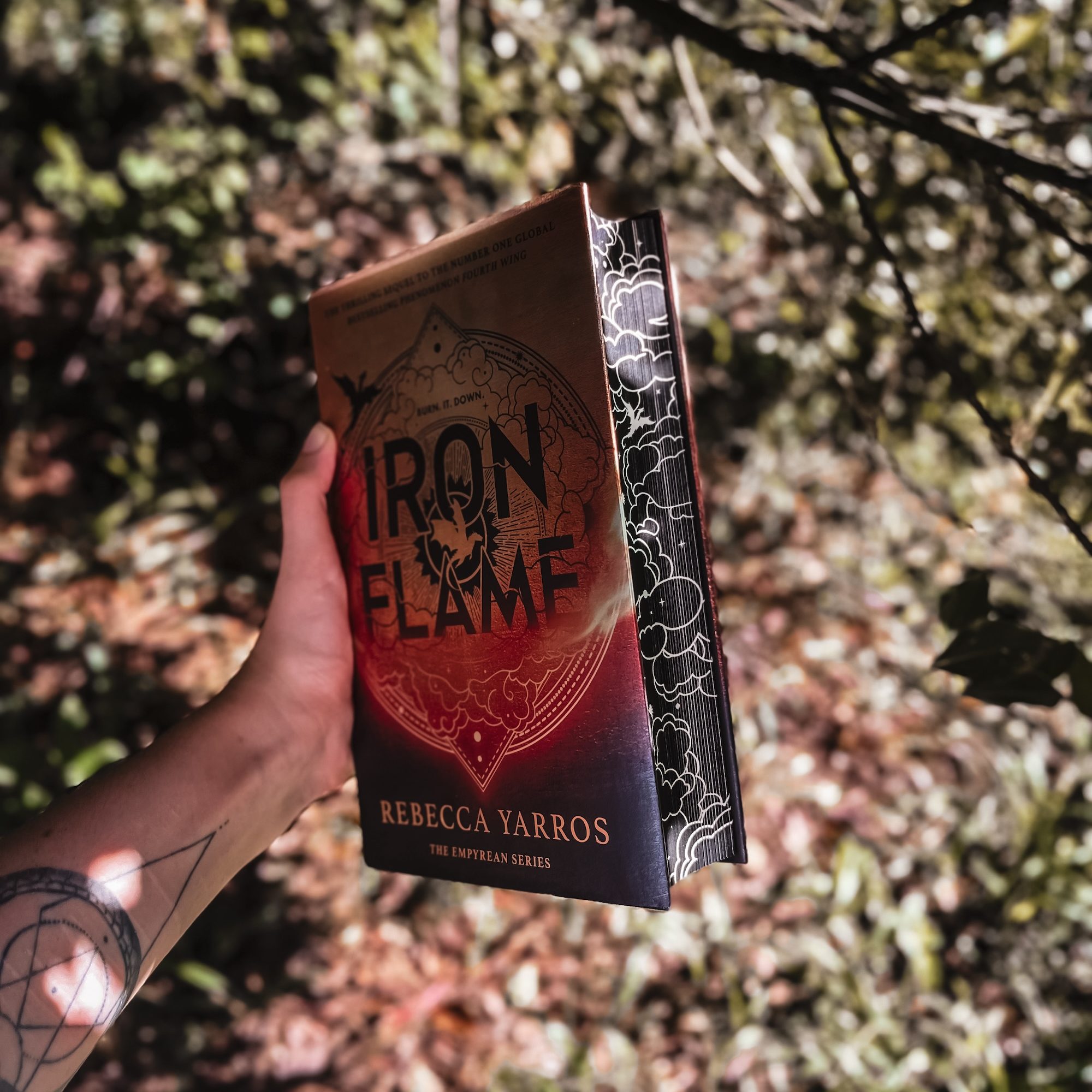 Hand holding Iron Flame by Rebecca Yarros in front of a forest.