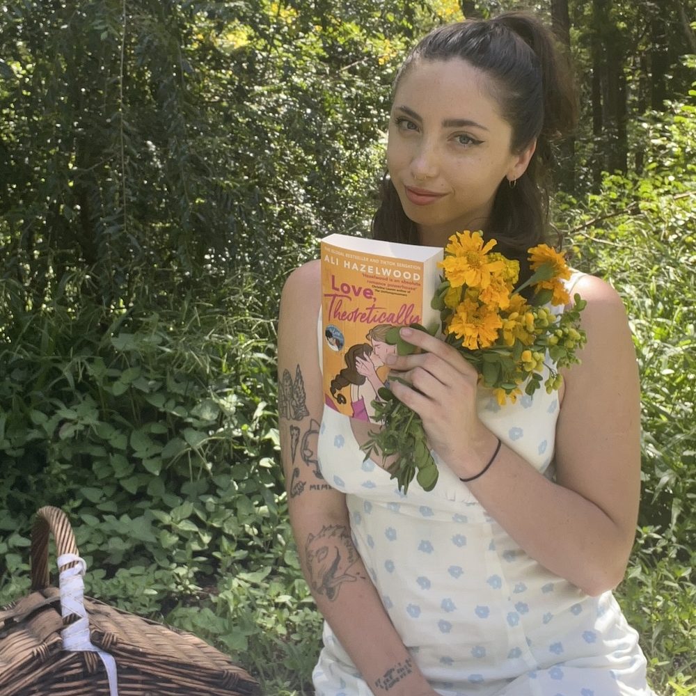 Ruby sitting in front of a forest and holding Love, Theoretically by Ali Hazelwood and some yellow flowers.