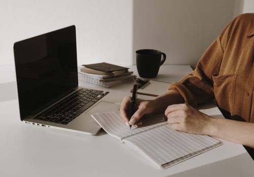 A woman writing on a notebook with a laptop beside her.