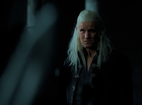 A man (Daemon) with long blonde hair looks off to the distance with tears in his eyes. He is wearing black.