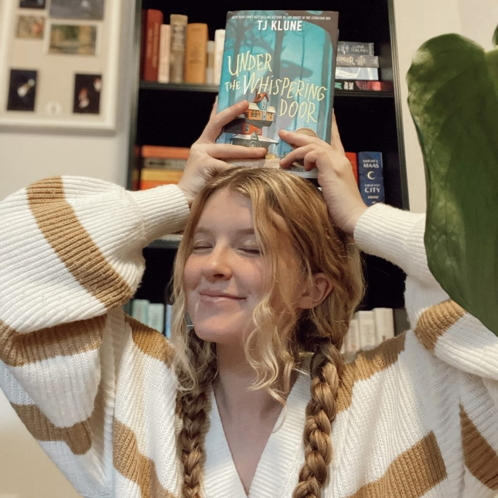 A smiling woman with reddish blonde hair holds a book above her head. The book is "Under the Whispering Door." She is wearing a tan and white striped sweater and has a bookshelf behind her.