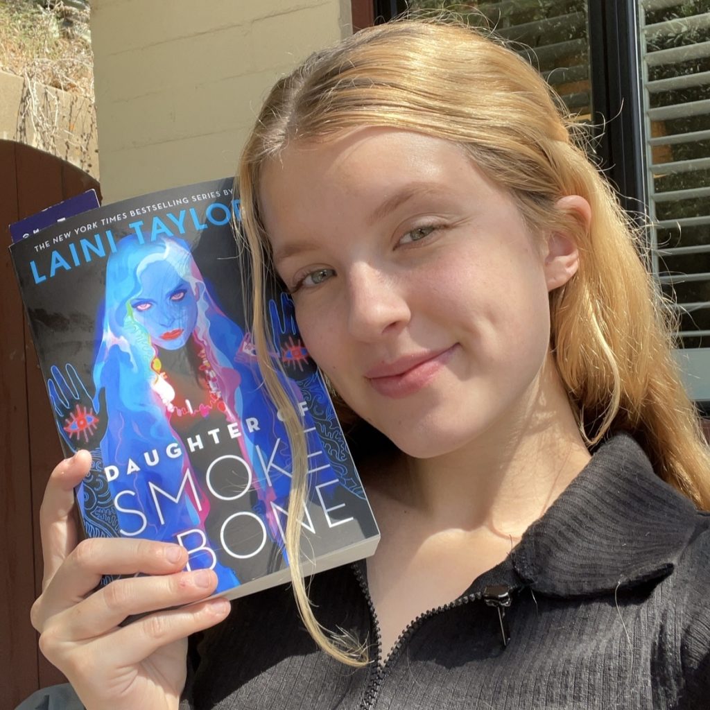 A woman with blonde hair and light skin smiles into the camera and holds up the book "Daughter of Smoke and Bone" next to her face. The image is from slightly below her shoulders up.