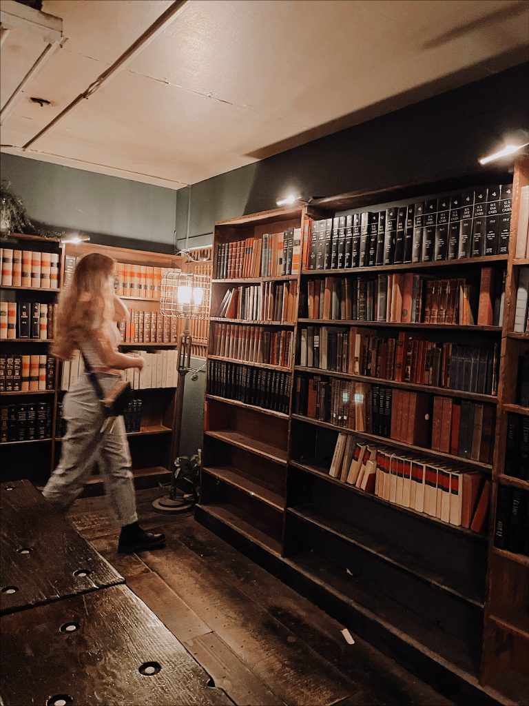 A blurry woman walks towards a dimly lit bookshelf. She is in a room with green walls and wooden floors and bookshelves.