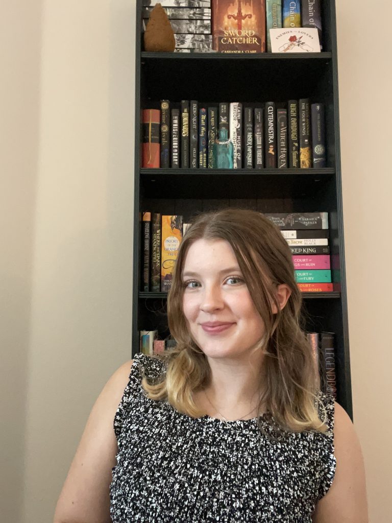 A woman with dirty blonde hair smiles at the camera. She is wearing a speckled black and white top and is sitting in front of a bookshelf.