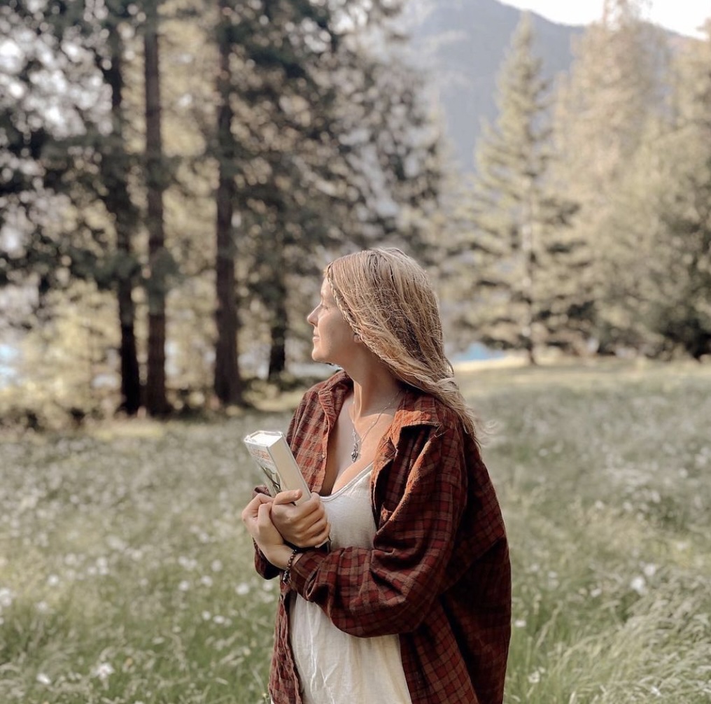 A woman with reddish blonde hair looks off to the left. She is wearing a red flannel over a white shirt and is holding a white book. She is standing in a green field with trees behind her.