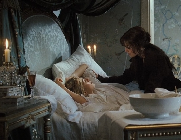 In an ornate blue bedroom, a woman with brown hair (Elizabeth) sits on a bed and leans slightly over a woman with blonde hair (Jane), who is laying down.