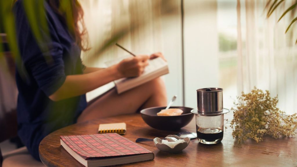 Woman writing in a journal in the background with a notebook and a water bottle on a table in the foreground.