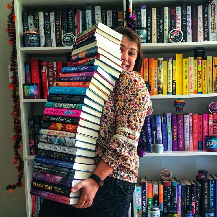 A brunette person holds a stack of books almost as tall as they are, with a smile, in front of a colorful bookshelf.