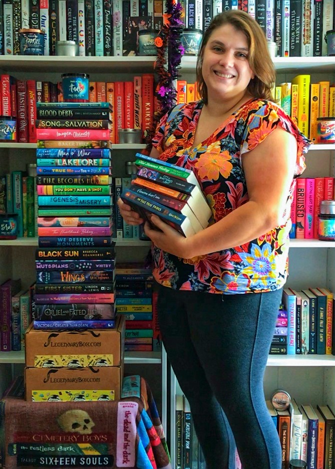 A brunette person holds a stack of books. There is also a large stack to their left. They are smiling and standing in front of a colorful bookshelf.