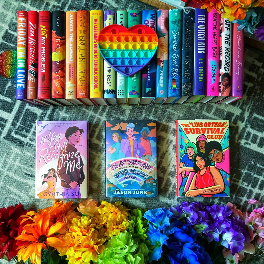 A line of colorful books, spine up, displayed on a carpet. Below them are three books, sitting cover up, including: "If You Still Recognize Me" by Cynthia So, "Riley Weaver Needs a Date to the Gaybutante Ball" by Jason June, and "The Luis Ortega Survival Club" by Sonora Reyes.