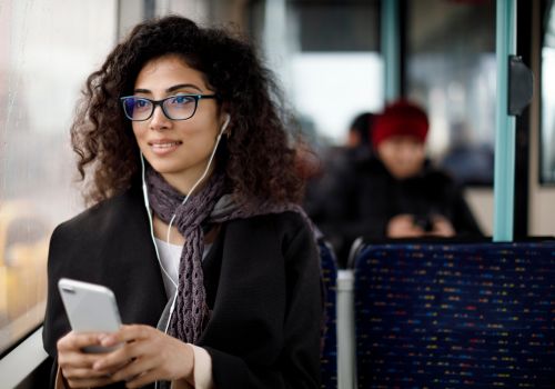 A woman on the bus wearing wired earphones, is holding a phone.