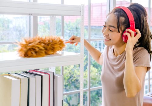 A smiling woman with headphones on is feather-dusting a set of shelves with books on it.