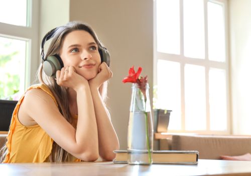 A woman with headphones on is sitting with her elbows on a desk and gazing into the distance. There is a book and flower in a vase on the desk.