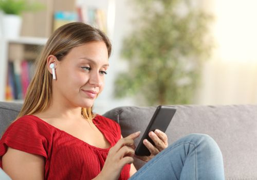 A smiling woman wearing earphones sits on a sofa, scrolling on her phone.