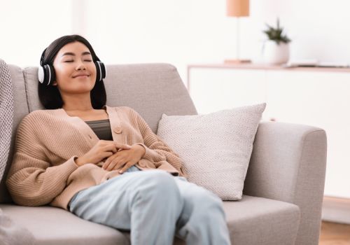 A woman sits slouched on a sofa smiling, with headphones on and her eyes closed.