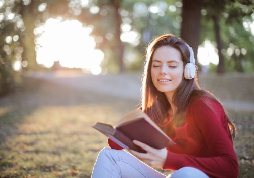 A woman wearing headphones is reading a book outside on a bright day. There is some foliage in the blurred background.