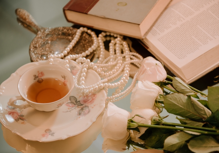 A teacup with tea in it on a saucer, small bouquet of white roses, an antique handheld mirror lying face-down, pearl necklace and pile of books are placed on a reflective surface.