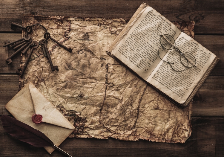 A crumpled vintage map on a wooden surface with a sealed letter and dark-coloured feather placed on its bottom left corner. A set of keys is placed on its top left corner and an open book with a pair of round glasses is placed on the top right corner of the map.