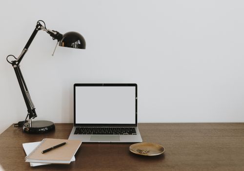 A black desklamp,, laptop with a blank white screen, short stack of notebooks with a pen on it and small dish with safety pins in it are placed on one side of a wooden desk.