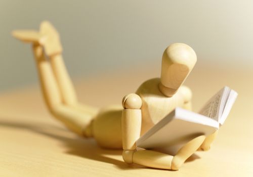 A wooden art model mannequin in a lying position with a book propped up in it's "hands" to look like it's reading. It's "legs" are raised and appear blurred in the background.