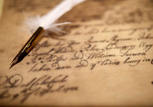 A blurred close-up shot of a quill pen lying on paper with writing on it in a fancy archaic-looking cursive style.