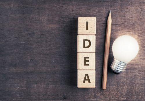 Four wooden blocks arranged vertically with letters on them that spell out "IDEA" are placed next to a pencil and light bulb on a dark brown wooden desk.
