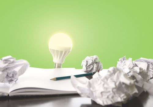 An open notebook, with a penil on it is surrounded by scrunched up pieces of paper, with a lightbulb and green background behind them.