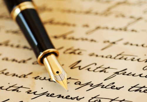 A fountain pen lies on top of a partially-blurred sheet of paper with cursive writing on it.