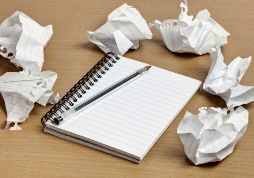 A light brown wooden surface with a black pen lying on an open notepad with six scrunched up pieces of paper placed around it.