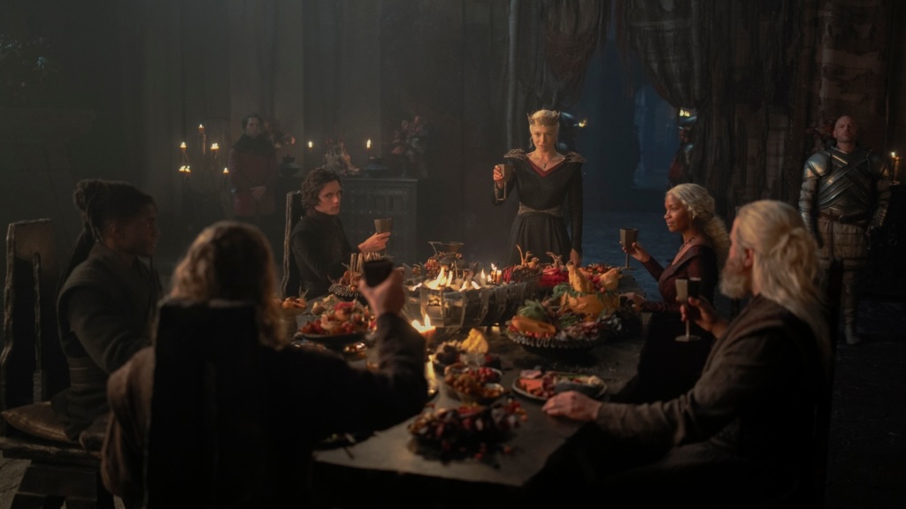 A variety of people sit at a long table with a ton of food on it. A woman with blonde hair and a crown (Rhaenyra) stands at the head, toasting.