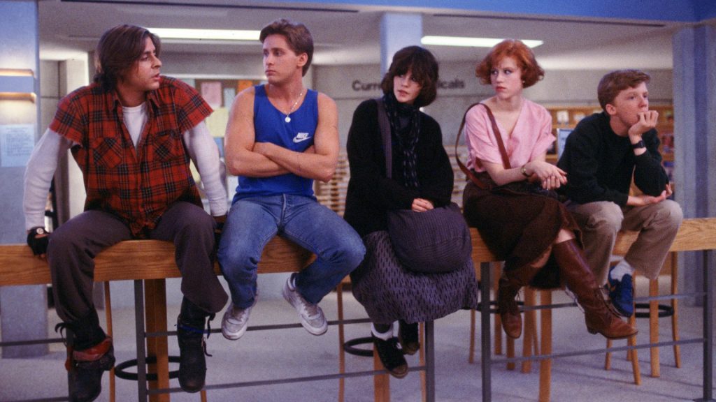 5 teenagers sit on a railing in a library. There are 3 boys, one with a red flannel, one with a blue tank top and one with a sweater and khakis. There are 2 girls, one all in black and the other with a pink shirt and brown skirt.