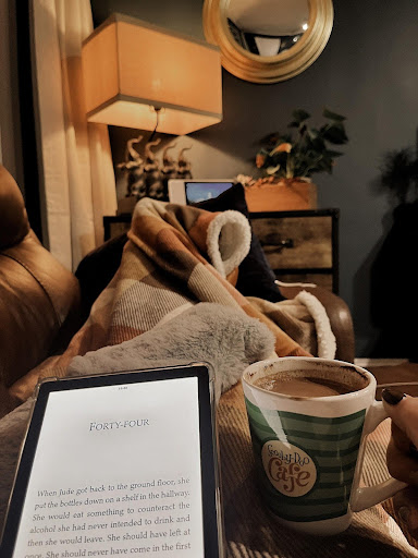 Image provided by bookstagrammer Laura of her on the couch, a blanket covering her legs as she holds a kindle and a drink in a mug.