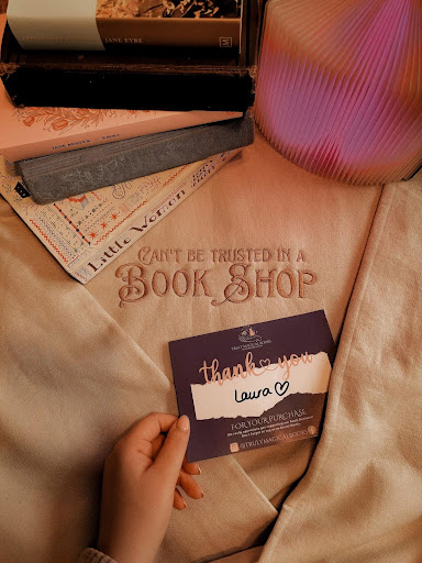 Image provided by bookstagrammer Laura of a cloth with "Can't be Trusted in a Books Shop" embraided on it. Laura is holding a card that says "thank you" with her named written on the card.