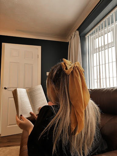 Image provided by bookstagrammer Laura of her backside facing the camera. She's holding an open book, half her hair tied up with a yellow bow.