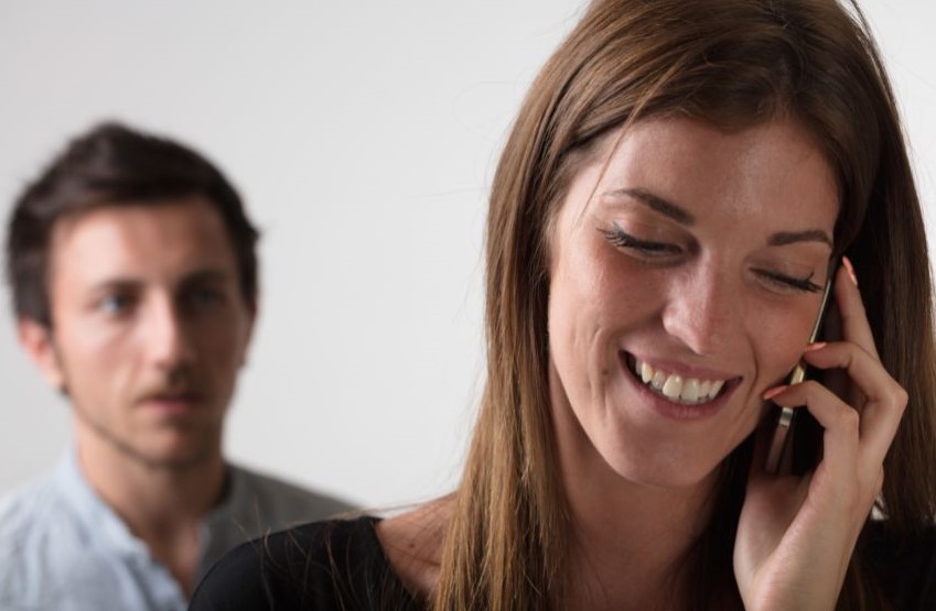 A woman smiling while on the phone and a man behind her looking angry