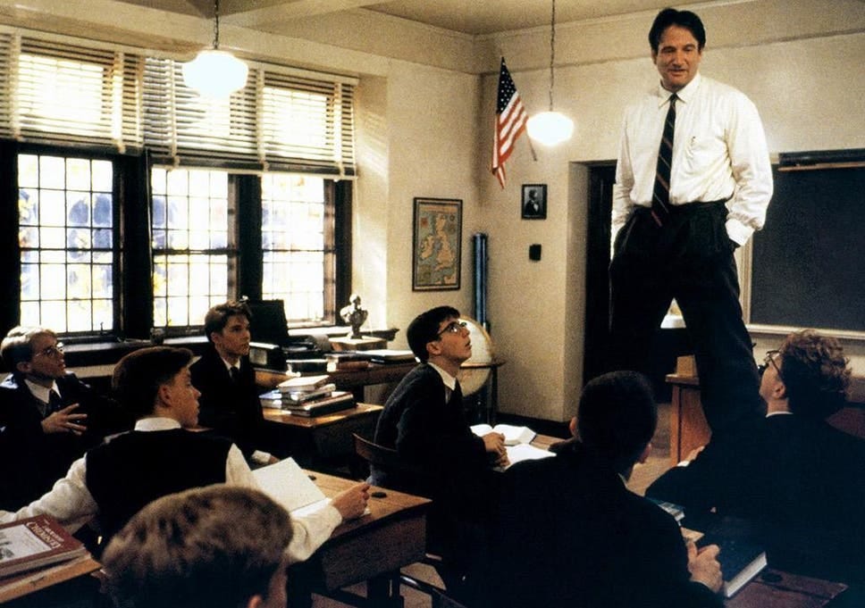 A still from the movie Dead Poets Society in which Robin Williams, playing a teacher, stands on top of a desk with his students gazing up at him.