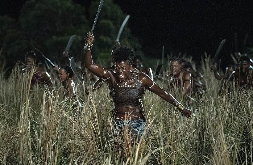 Viola Davis leads a group of female African warriors to war. They are running through tall grass and brandishing swords over their heads.