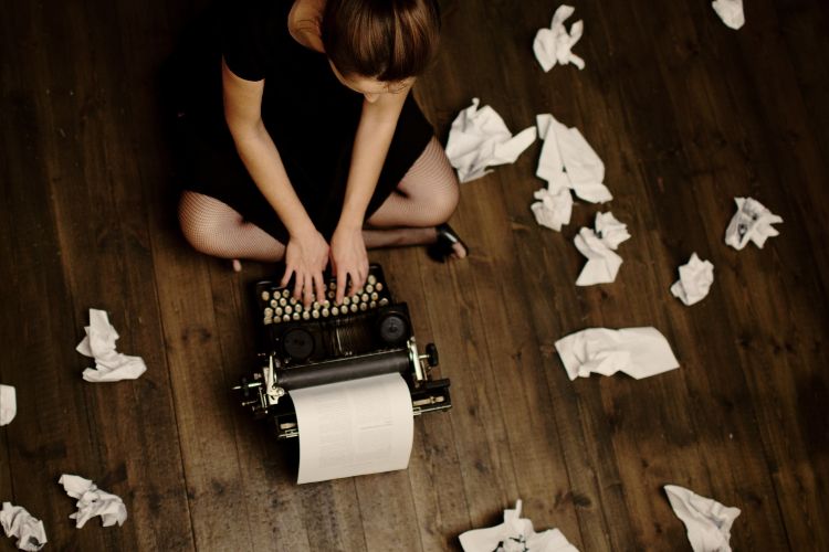 A woman writes on a typewriter on the floor, surrounded by crumbled pieces of paper.
