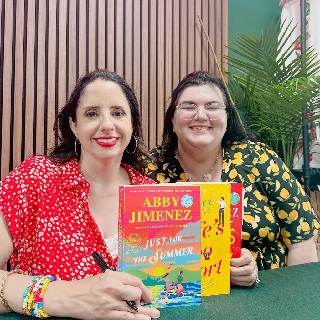 A woman (Abby Jimenez) with red lipstick and a red blouse holds up three books she has written. Next to her is another woman (Bree) with short brown hair and a blouse with lemons. They are both smiling at the camera.